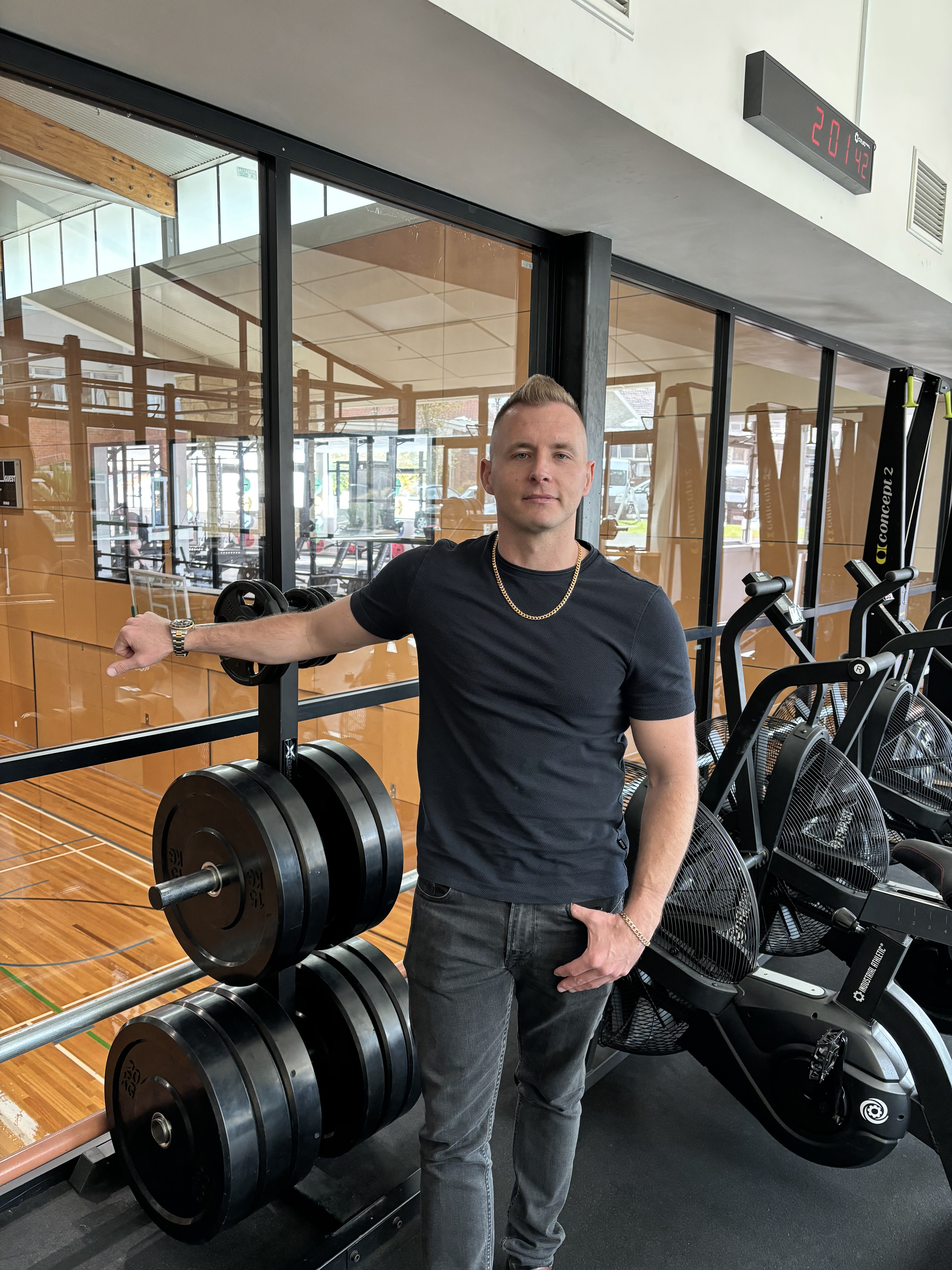 Man in his 30s standing with his hand on a weight in a gym. 