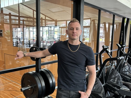 Man in his 30s standing with his hand on a weight in a gym. 