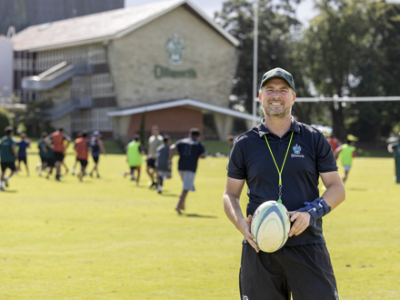 Director of Rugby holding a rugby ball with student in the background 