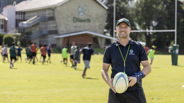 Director of Rugby holding a rugby ball with student in the background 